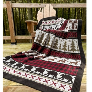 Peut inclure: Une couverture en crochet marron, rouge et blanc avec un motif de forêt comprenant des ours, des arbres et des cerfs. La couverture est drapé sur une chaise en bois sur une terrasse en bois.