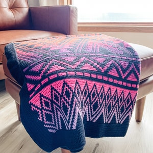 May include: A black and pink crocheted throw blanket with a geometric pattern. The blanket is draped over a brown leather ottoman.