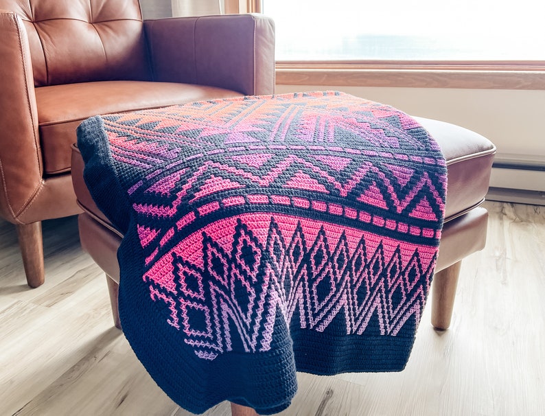 May include: A black and pink crocheted throw blanket with a geometric pattern. The blanket is draped over a brown leather ottoman.