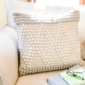 May include: A cream-colored crocheted pillow with a textured, raised pattern. The pillow is sitting on a light beige couch with a stack of books and glasses on top.