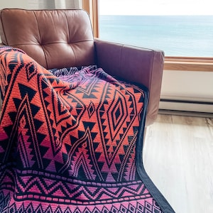 May include: A colourful crocheted throw blanket with a geometric pattern in shades of pink, orange, and black. The blanket is draped over a brown leather armchair in front of a window with a view of the sea.