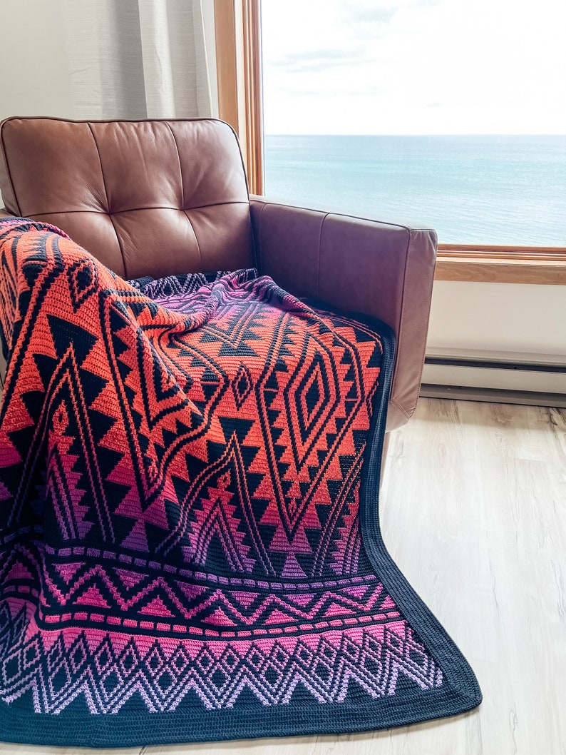 May include: A colourful crocheted throw blanket with a geometric pattern in shades of pink, orange, and black. The blanket is draped over a brown leather armchair in front of a window with a view of the sea.