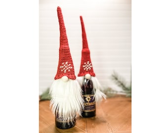 May include: Two red crocheted gnome bottle toppers with white fur beards and snowflake designs. The gnomes are on top of wine bottles.