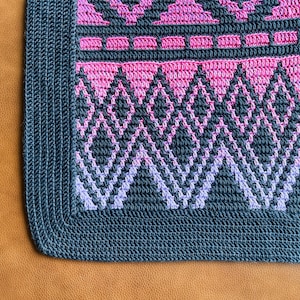 May include: A close-up of a crocheted blanket with a geometric pattern in shades of pink, purple, and grey. The blanket has a dark grey border.