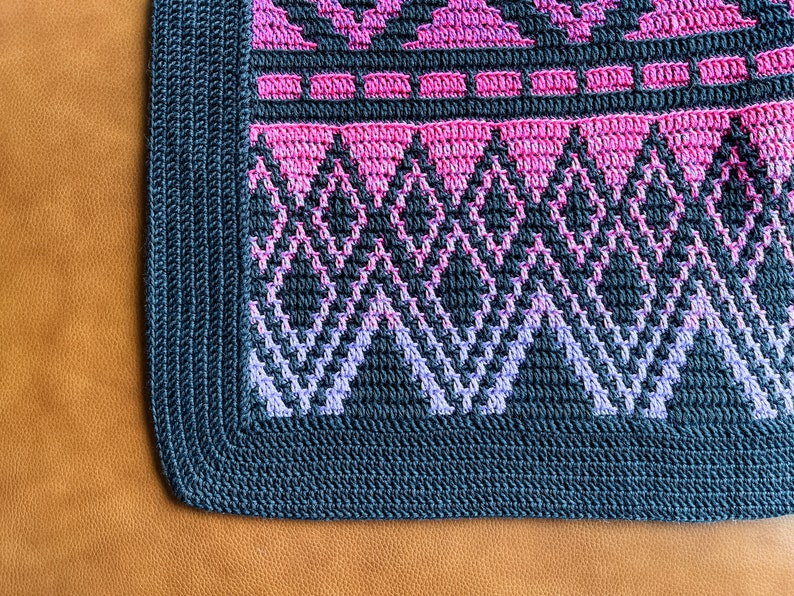 May include: A close-up of a crocheted blanket with a geometric pattern in shades of pink, purple, and grey. The blanket has a dark grey border.
