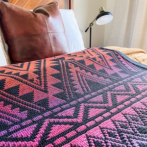 May include: A close-up of a colourful crocheted throw blanket with a geometric pattern in shades of pink, purple, and black. The blanket is draped over a bed with a brown leather pillow.