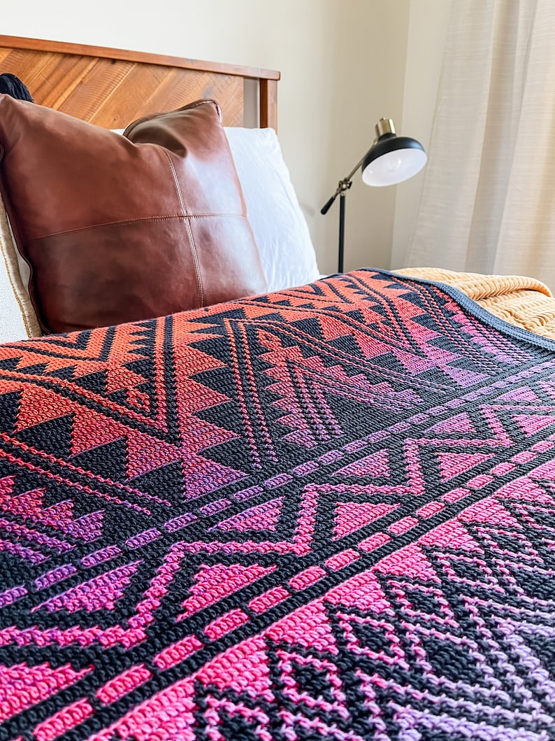 May include: A close-up of a colourful crocheted throw blanket with a geometric pattern in shades of pink, purple, and black. The blanket is draped over a bed with a brown leather pillow.