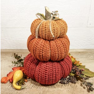 Op de afbeelding: Een stapel van drie gehaakte pompoenen in oranje en rode tinten, bekroond met een takje en touw. Decoratieve kalebassen en herfstbladeren omringen de pompoenen, wat een seizoensgebonden herfstdisplay creëert.