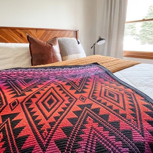 May include: A bed with a wooden headboard and a colourful geometric patterned blanket. The blanket has shades of pink, orange, and black. The bed is made with white sheets and pillows.