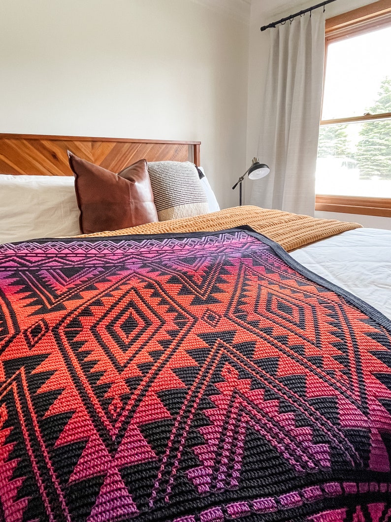 May include: A bed with a wooden headboard and a colourful geometric patterned blanket. The blanket has shades of pink, orange, and black. The bed is made with white sheets and pillows.