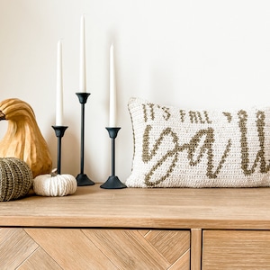 May include: A white crocheted pillow with the text "It's Fall Y'all" in brown yarn. The pillow is sitting on a wooden shelf with three black candle holders and three white and brown gourds.
