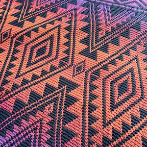 May include: A close-up of a crocheted blanket with a geometric pattern in shades of pink, orange, and black.
