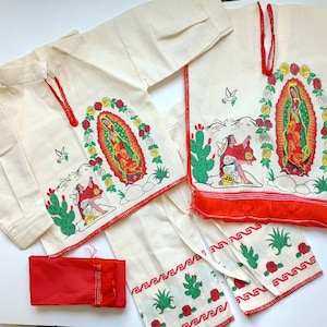 May include: A white cotton outfit for a child, featuring a long-sleeved shirt and pants. The shirt and pants have embroidered images of the Virgin Mary, roses, and cacti. The outfit also includes a red sash with fringe.