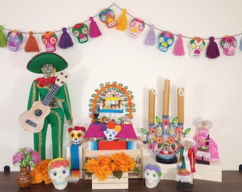 May include: A Day of the Dead display featuring a garland of colourful skull ornaments, a green mariachi figure with a guitar, a tiered stand, a three-candle candelabra, two small dolls, and several decorative sugar skulls with floral accents.