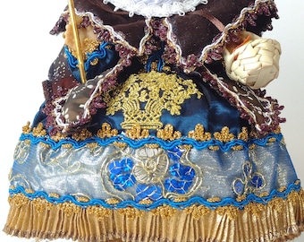 Puede incluir: Una pequeña figurita de porcelana de un niño vestido con una túnica azul y marrón con ribetes dorados. El niño lleva un sombrero de paja y sostiene un bastón. La figurita está sentada sobre una base de madera.