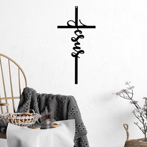 The word Jesus is written vertically in a casual, cursive font and connected as part of an actual cross to hang on the wall. The sign is shown with a chair and potted flowers.