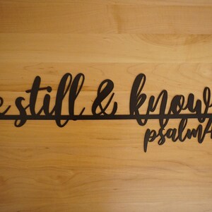 The words be still & know with the scriptural reference are displayed in a single cursive font, lower case lettering, and connected together to hang on the wall as a single piece. Sign is black on a wooden background.