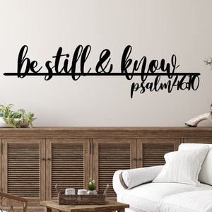 The words be still & know with the scriptural reference are displayed in a single cursive font, lower case lettering, and connected together to hang on a wall as a single piece. The background wall is white with sign hanging above a wooden cabinet.