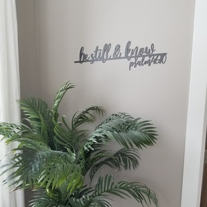 The words be still & know with the scriptural reference are displayed in a single cursive font, lower case lettering, and connected together to hang on the wall as a single piece. Sign is black hanging above green plant.