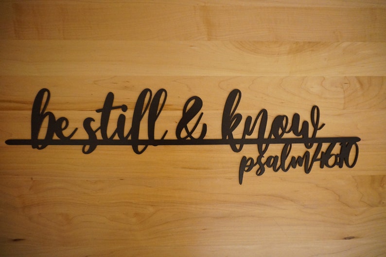 The words be still & know with the scriptural reference are displayed in a single cursive font, lower case lettering, and connected together to hang on the wall as a single piece. Sign is black on a wooden background.