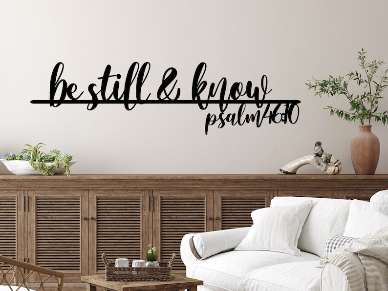 The words be still & know with the scriptural reference are displayed in a single cursive font, lower case lettering, and connected together to hang on a wall as a single piece. The background wall is white with sign hanging above a wooden cabinet.