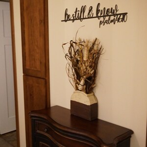 The words be still & know with the scriptural reference are displayed in a single cursive font, lower case lettering, and connected together to hang on the wall as a single piece. Sign is black hanging above cabinet with dried flowers.