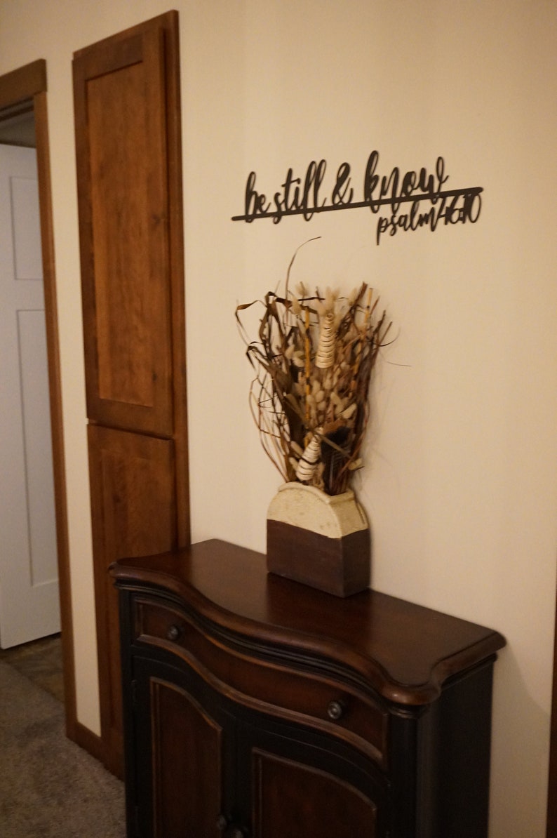 The words be still & know with the scriptural reference are displayed in a single cursive font, lower case lettering, and connected together to hang on the wall as a single piece. Sign is black hanging above cabinet with dried flowers.
