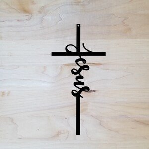 The word Jesus is written vertically in a casual, cursive font and connected as part of an actual cross to hang on the wall. The sign is shown with a wooden background.