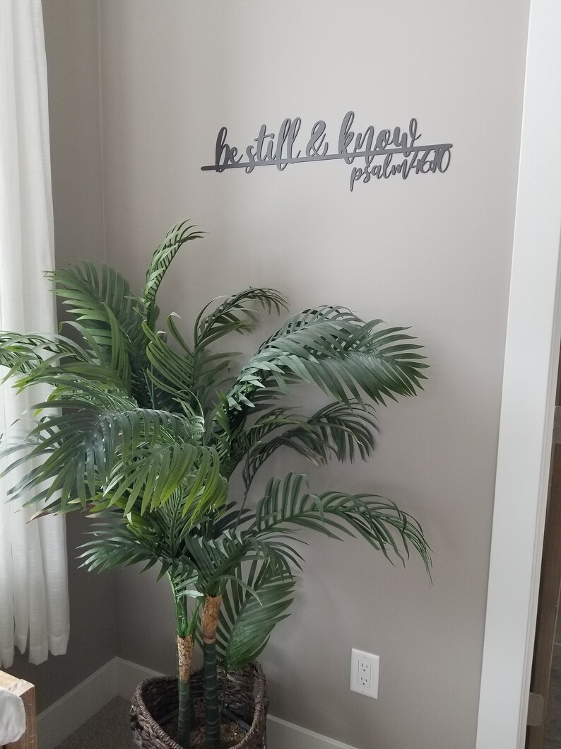 The words be still & know with the scriptural reference are displayed in a single cursive font, lower case lettering, and connected together to hang on the wall as a single piece. Sign is black hanging above green plant.