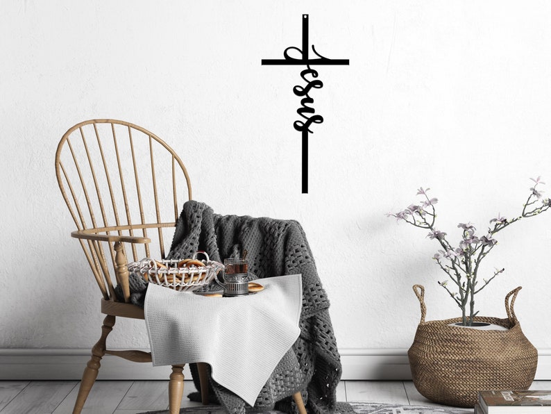 The word Jesus is written vertically in a casual, cursive font and connected as part of an actual cross to hang on the wall. The sign is shown with a chair and potted flowers.