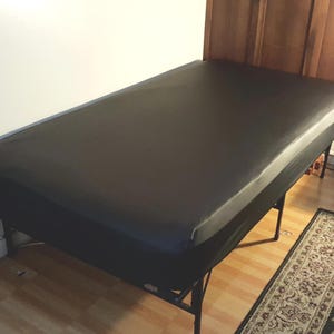 May include: A black massage table with a rectangular shape. The table has a black surface and is supported by a black metal frame. The table is placed against a white wall and a wooden headboard.