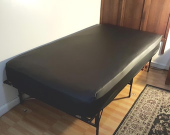 May include: A black massage table with a rectangular shape. The table has a black surface and is supported by a black metal frame. The table is placed against a white wall and a wooden headboard.