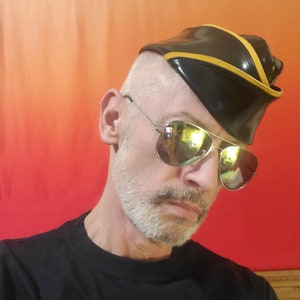May include: A man wearing a black leather hat with a yellow stripe and aviator sunglasses. The hat has a short brim and is angled on his head. He is wearing a black t-shirt.