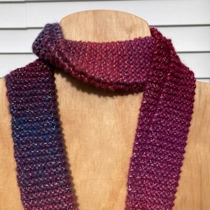 May include: A knitted scarf in shades of purple, pink, and blue. The scarf is made with a sparkly yarn and has a subtle ombre effect.