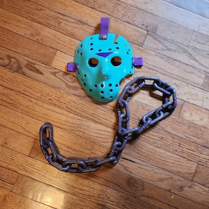 Puede incluir: Una máscara de hockey turquesa con correas moradas y un detalle triangular morado a juego. Una cadena gris oscuro está al lado de la máscara. La máscara tiene varios agujeros pequeños y una gran abertura en el lado derecho.