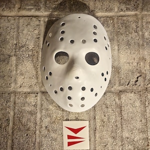 Könnte beinhalten: Eine weiße Hockeymaske mit zahlreichen kleinen, gleichmäßig verteilten Löchern und großen Augenöffnungen. Darunter ein kleiner Aufkleber mit einem roten Pfeildesign. Die Maske ist eine Requisite aus einem Horrorfilm.