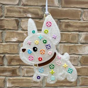 May include: A translucent white French bulldog ornament featuring a glittery finish and colorful, small floral and star-shaped accents across its body. The dog has a brown collar and is suspended by a thin white string attached to a gold-toned ring.