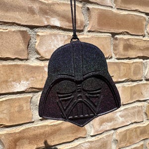 May include: A glittery, black and purple Darth Vader helmet-shaped ornament hanging against a brick wall. The ornament has a detailed design of the character's mask and is suspended by a black cord. The brick wall is a mix of brown and tan bricks.