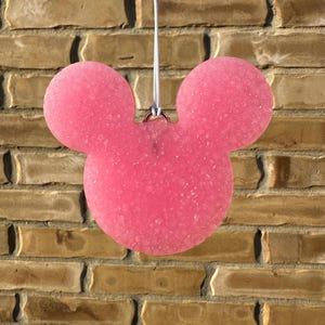May include: A pink, Mickey Mouse-shaped air freshener hanging against a brick wall. The air freshener is a solid pink color with a textured surface and a small silver ring for hanging. A white string is attached to the ring.