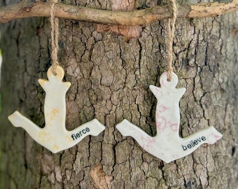 May include: Two ceramic anchors hanging from a tree branch. The anchor on the left is yellow with the word "fierce" written on it. The anchor on the right is pink with the word "believe" written on it.