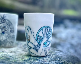 May include: Two white ceramic mugs with blue and white illustrations. One mug features a woman holding a spinning wheel, the other mug features a woman with a floral design.