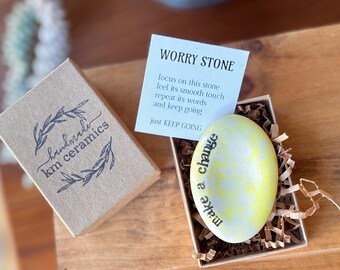 May include: A small yellow worry stone with the words "make a change" inscribed on it. The stone is in a small box with a card that reads "Worry Stone, focus on this stone, feel its smooth touch, repeat its words and keep going, just KEEP GOING."