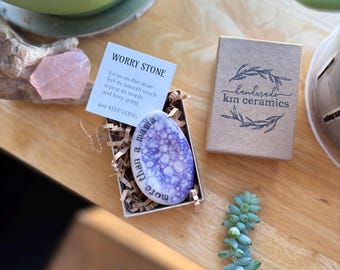 May include: A small, purple and white worry stone with the text "More than a mama" on it. The stone is in a small box with brown paper shreds. The box also has a card that says "Worry Stone" and instructions on how to use it. The box is on a wooden surface with a green plant in the background.