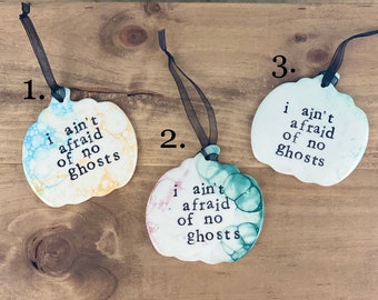 May include: Three white ceramic pumpkin ornaments with black text that reads "i ain't afraid of no ghosts". Each ornament has a black ribbon for hanging.