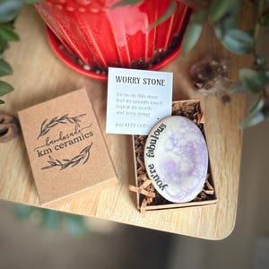 Può includere: Una piccola pietra di preoccupazione di colore viola e bianco con il testo "You're fabulous" scritto su di essa. La pietra è in una piccola scatola con un biglietto che dice "Worry Stone" e "Just keep going".