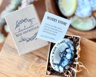 May include: A small, white ceramic worry stone with black ink lettering that reads "O Face Your Fears". The stone is in a small box with brown paper shreds. A card with the text "Worry Stone" and instructions for using the stone is visible.