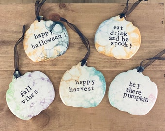 May include: Five ceramic pumpkin ornaments with black painted text. The ornaments are decorated with a marbled paint effect in shades of white, green, blue, and yellow. The text on the ornaments reads: "happy halloween!" "eat drink and be spooky" "fall vibes" "happy harvest" "hey there pumpkin"