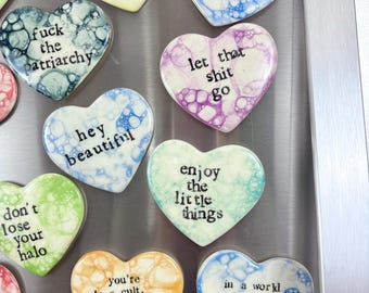 May include: A collection of heart-shaped magnets with various colors and phrases. The magnets feature quotes like "I'm all peace, love, light," "Let that shit go," and "In a world full of hate, be love." The magnets are displayed on a metallic surface.