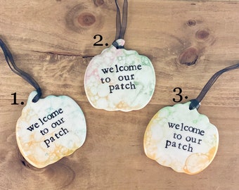 May include: Three ceramic pumpkin ornaments with black ribbon hangers. Each pumpkin has a different watercolor-like design and the text "welcome to our patch" in black lettering.