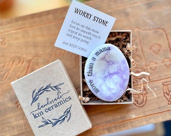 May include: A purple worry stone with the text "more than a mama" inscribed on it. The stone is in a small box with a brown paper label that says "handmade km ceramics".